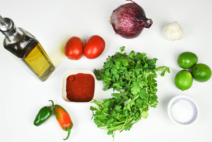 Chimichurri Rojo ingredients including olive oil, roma tomatoes, red onion, garlic, limes, cilantro, jalapenos, and seasonings