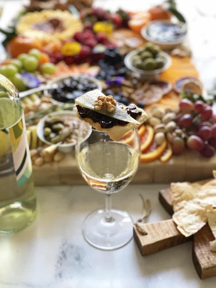 An epic cheeseboard with a glass of white wine in front that has crackers topped with Pinot Prune Jam, cheese, and nuts.