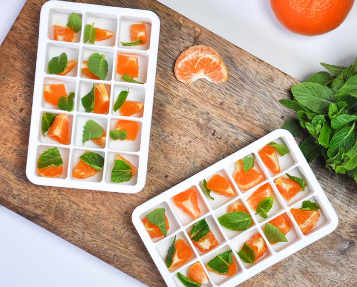 Two ice cube trays with a piece of citrus and mint in each ice cube square 