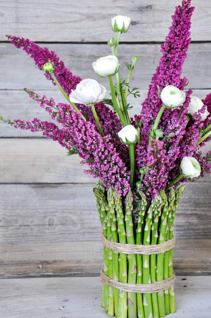 Out of the Box Decorating Ideas for Spring using fresh asparagus. A cylinder vase is covered with fresh asparagus and tied with twine. A bouquet of flowers is in the vase.