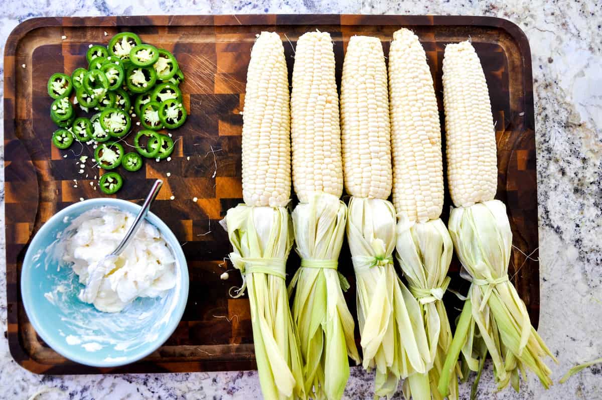 Husk peeled down the corn husk on a cutting board next to chopped jalapenos and cream cheese. 