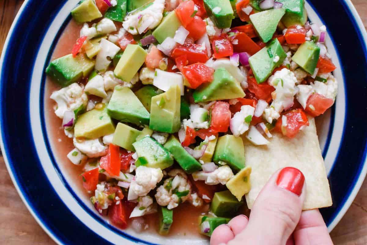 This vegan ceviche is featured in a roundup of California avocados recipes. It is a bowl full of minced vegetable sin red, white and green rtossed in a flavorful dressing and being scooped with a chip, 