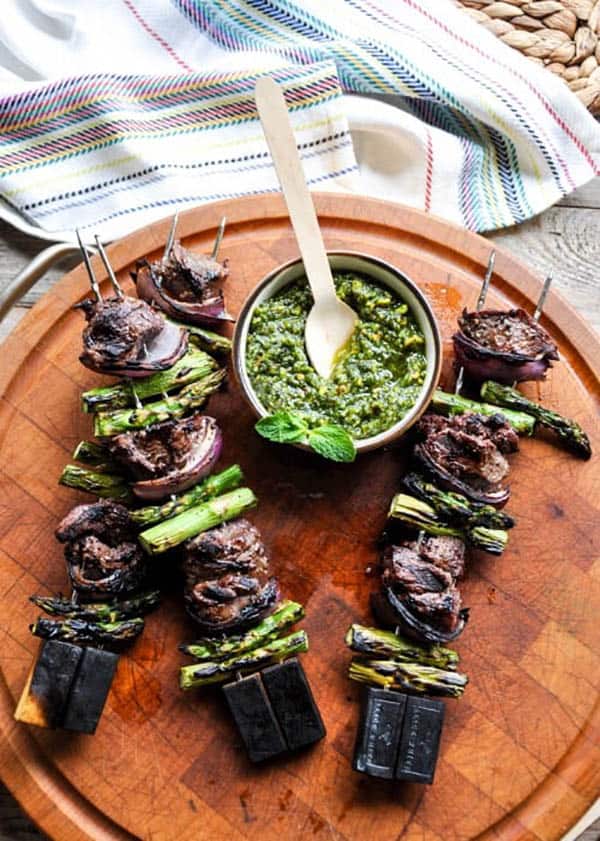 3 grilled lamb asparagus and red onion kabobs on a wooden platter with a bowl of pistachio mint pesto.