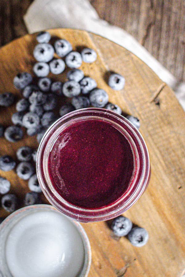 A jar of homemade Blueberry Jalapeño BBQ sauce is on a wooden board next to a jar of honey and fresh blueberries. 