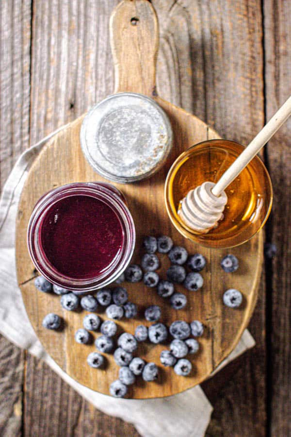 A jar of homemade Blueberry Jalapeño BBQ sauce is on a wooden board next to a jar of honey and fresh blueberries.