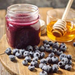 A jar of homemade Blueberry Jalapeño BBQ sauce is on a wooden board next to a jar of honey and fresh blueberries.