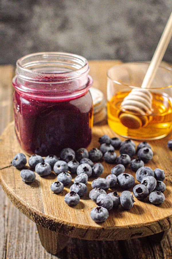 A jar of homemade Blueberry Jalapeño BBQ sauce is on a wooden board next to a jar of honey and fresh blueberries.