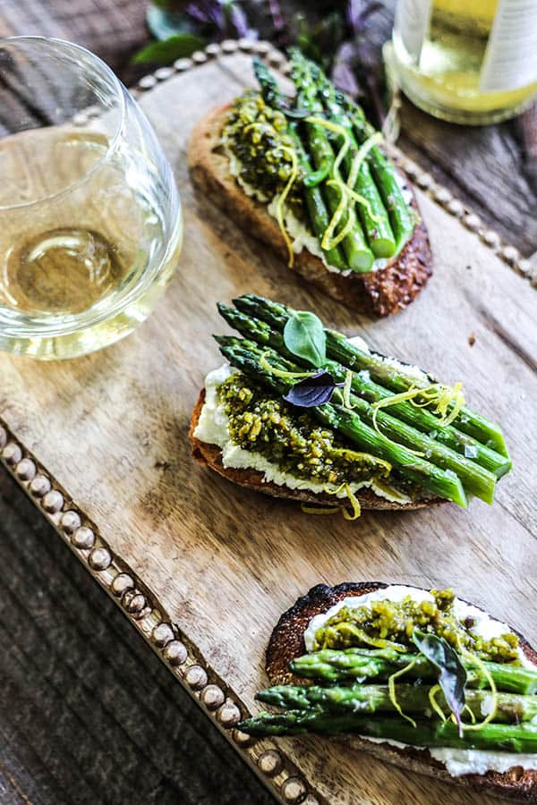 3 bruschetta with Warm Goat Cheese, Roasted Asparagus, and Pistachio Pesto on a wooden serving board next to a glass of white wine.