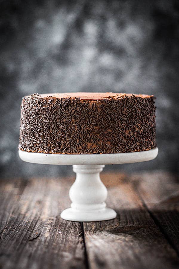 A chocolate cake that is made with prunes on a white cake stand. The cake has chocolate icing and chocolate sprinkles coat the sides.