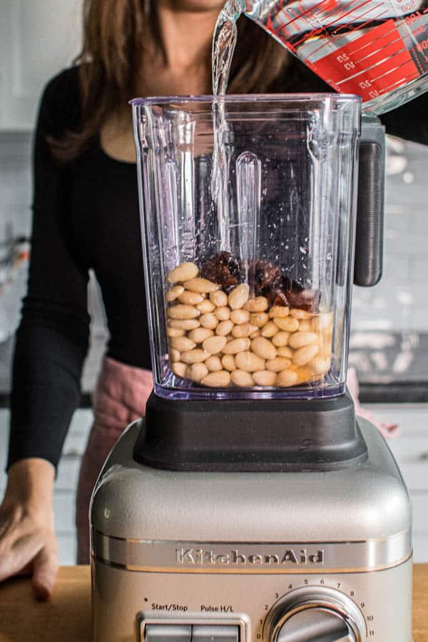 A woman pouring filtered water into a blender carafe with soaked blanche almonds and soaked diced prunes. This is how to make almond milk at home.