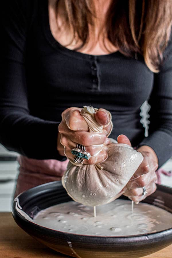 This is how to make almond milk! Blended almonds, prunes and filtered water mixture is being stained through a nut milk bag into a large bowl. A woman is gently squeezing the bad to extract all of the almond milk. 