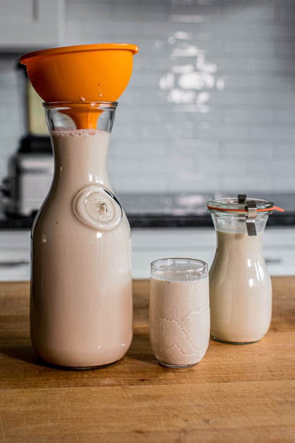 Almond milk being funneled into a large carafe on a counter next to a glass of prepared almond milk and a smaller carafe of almond milk creamer. 