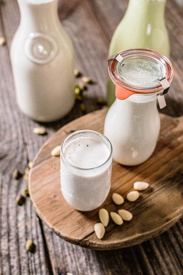 Multiple carafes of almond milk, pistachio milk, almond coffee creamer, and a glass of prepared vanilla almond milk are all on a table together showing how to store almond milk in glass carafes or jars. 