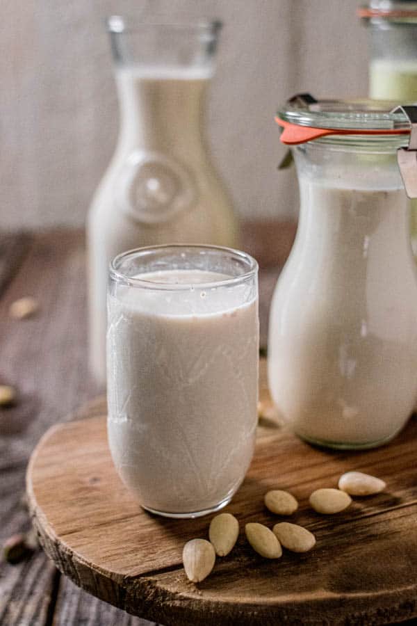 A glass of homemade almond milk sits next to a small jar of homemade almond milk coffee creamer and a large carafe of almond milk.