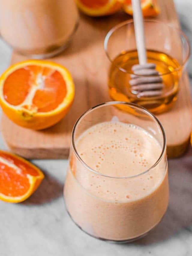 Two orange julius smoothies on a marble counter top next to a small jar of honey and Cara Cara orange slices.