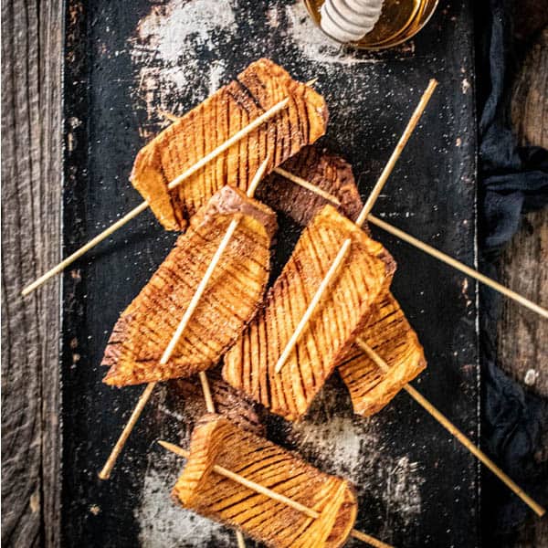 5 Hasselback sweet potato skewers next to a jar of honey on a serving tray.