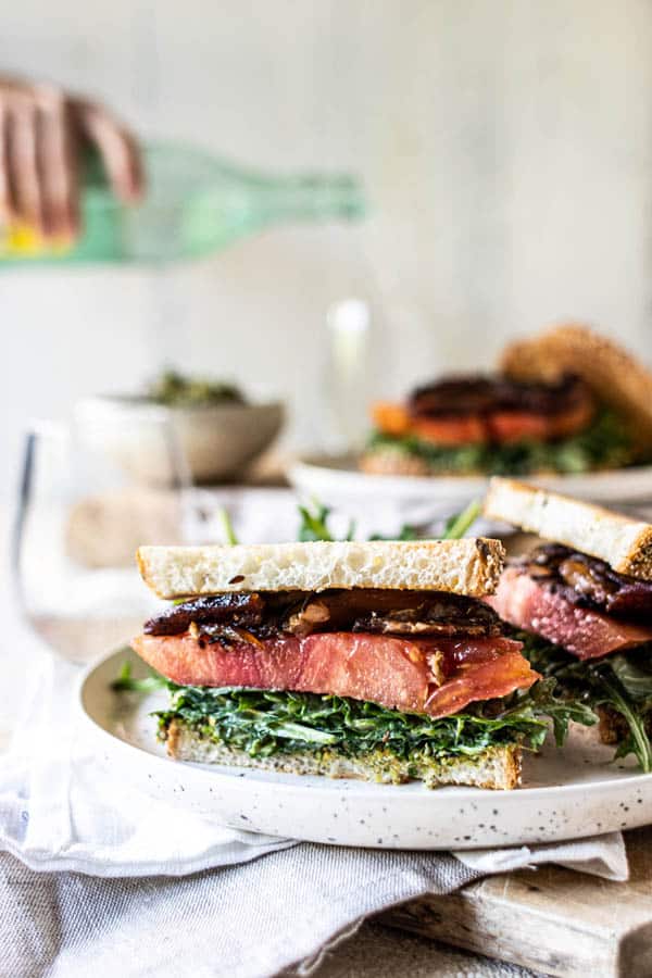 A halved Ultimate California Grown BLT sandwich on a plate with an arugula salad. A woman is pouring a glass of sparkling water in the background.