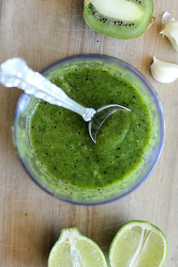 A bowl of kiwi salsa verde on a table next to kiwis and limes