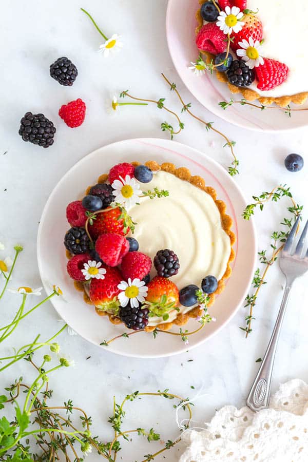 Two individual tarts on serving plates. The tarts have a beautiful white filling and are topped with red, white and blue berries as well as chamomile flowers. 