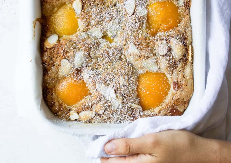 A cake made in a white baking dish that is rectangular. the cake is topped with almond slivers and powdered sugar. There are canned peach halves peeking up from the batter.