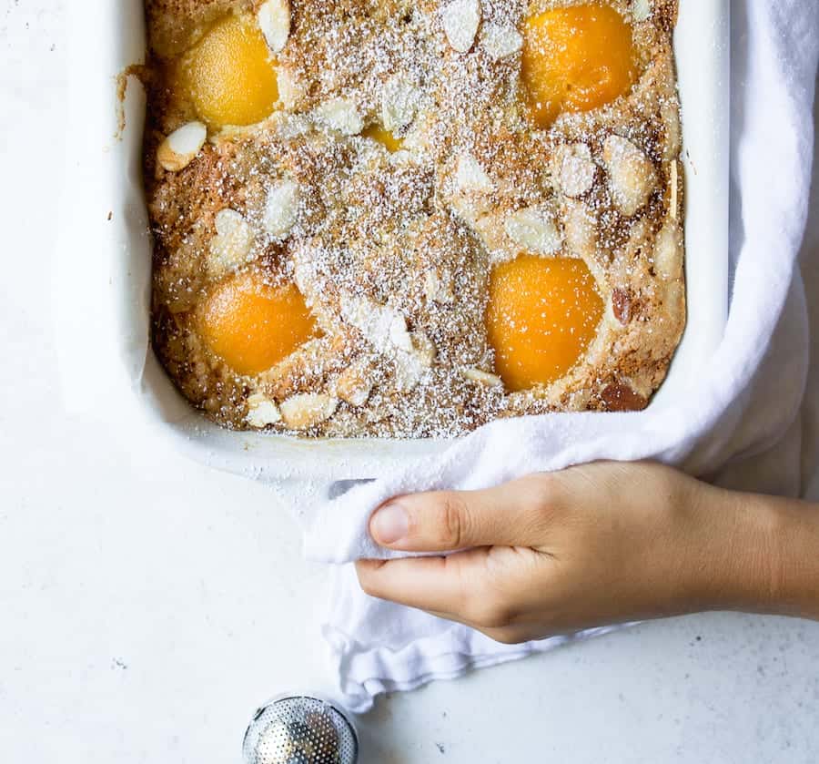 A cake made in a white baking dish that is rectangular. the cake is topped with almond slivers and powdered sugar. There are canned peach halves peeking up from the batter. 