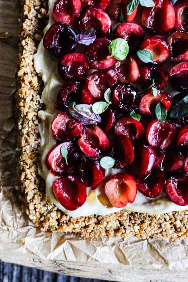 A tart with a pretzel crust is topped with a white cream cheese layer and fresh cherry halves then topped with basil.