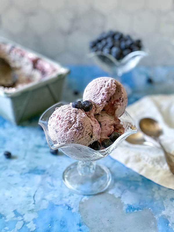 Three scoops of fresh blueberry ice cream in a fluted serving dish topped with fresh blueberries. 