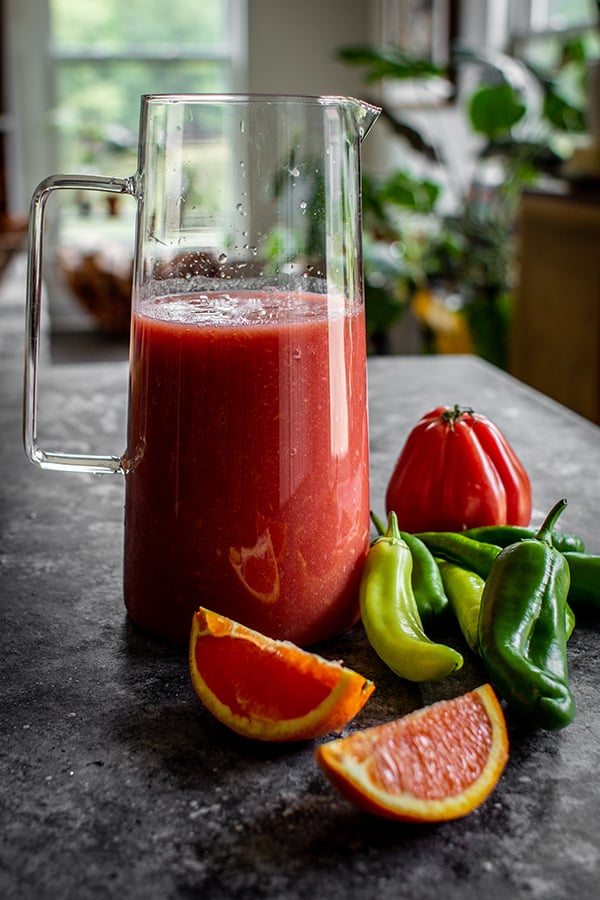 A pitcher of homemade Bloody Mary mix next to fresh orange slices, peppers and an heirloom tomato on a table.