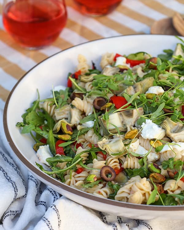 A bowl of pasta salad with black olives, arugula, artichokes and goat cheese. 