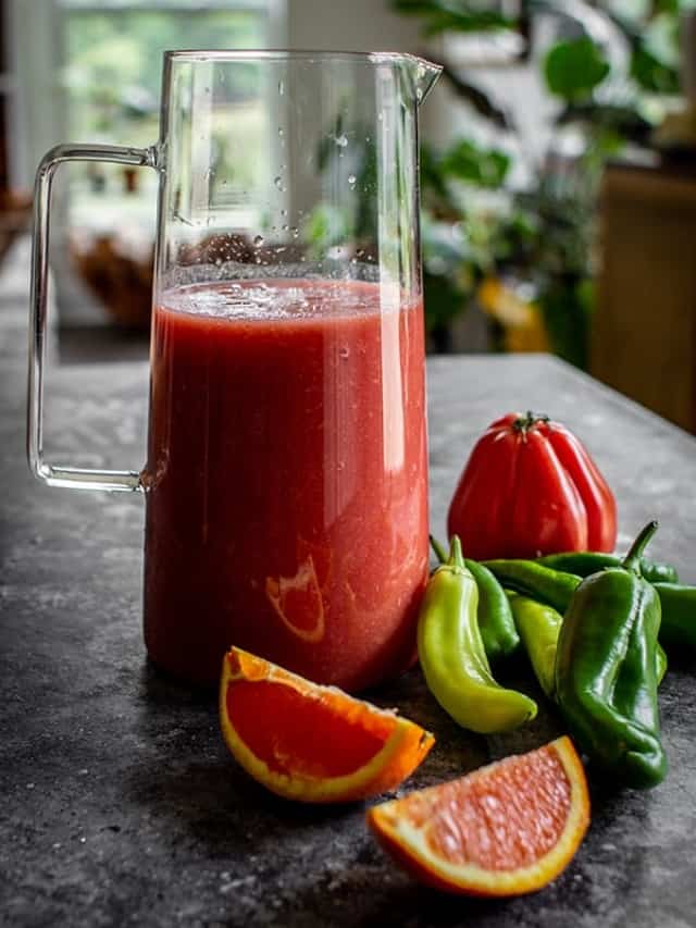 A pitcher of homemade Bloody Mary mix next to fresh orange slices, peppers and an heirloom tomato on a table.