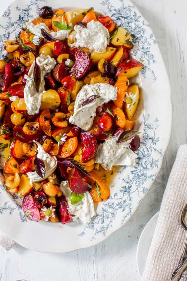 A large white platter with blue trim is filled with chopped apricots, cherries, plums, and peaches then topped with burrata, basil, and chamomile.