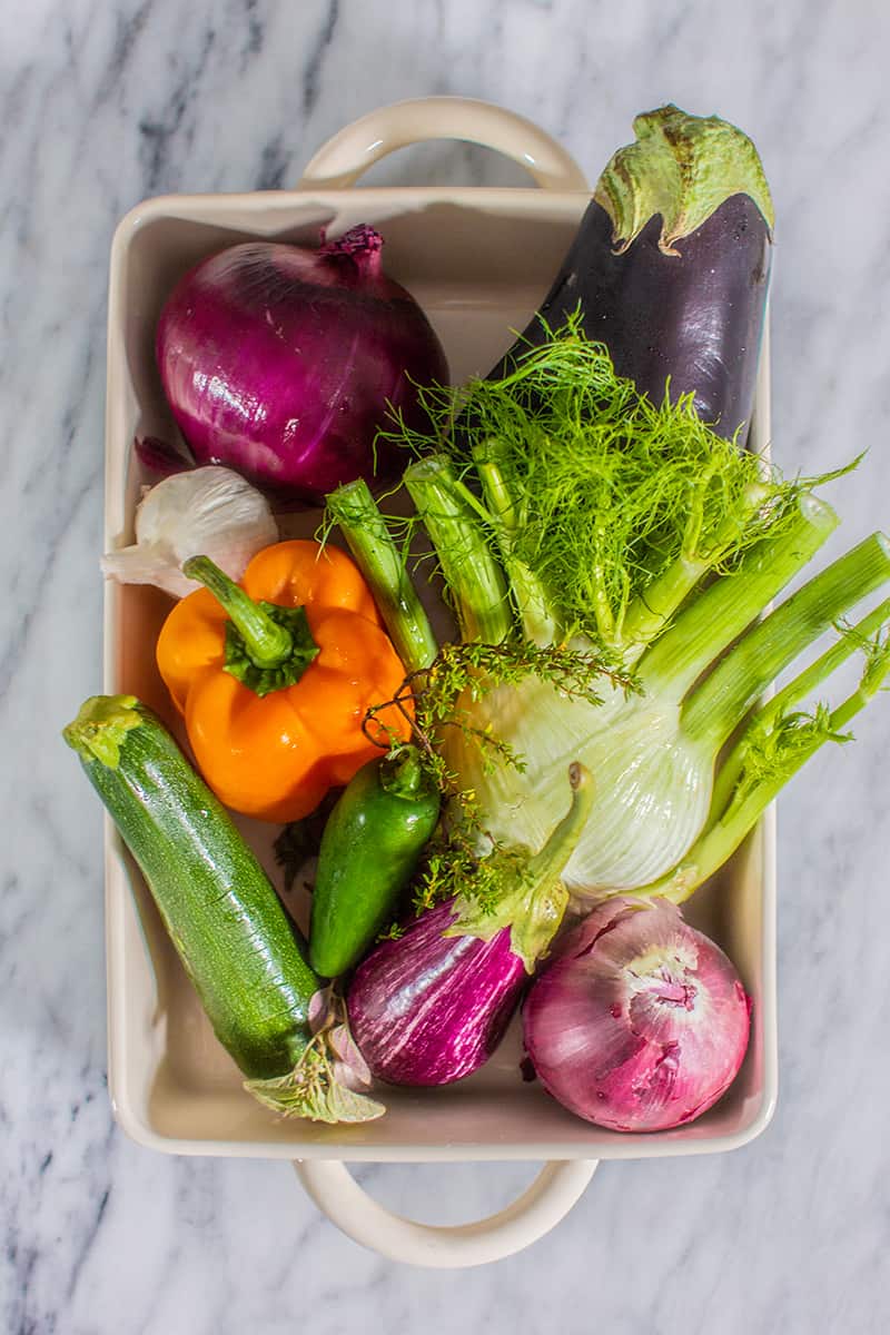 A variety of fresh vegetables to make caponata of eggplant in a roasting dish. There is fennel, eggplant, peppers, zucchini, onions and garlic.