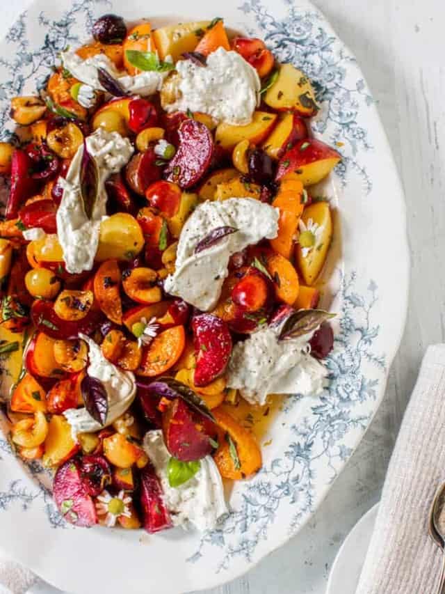 A large white platter with blue trim is filled with chopped apricots, cherries, plums, and peaches then topped with burrata, basil, and chamomile.