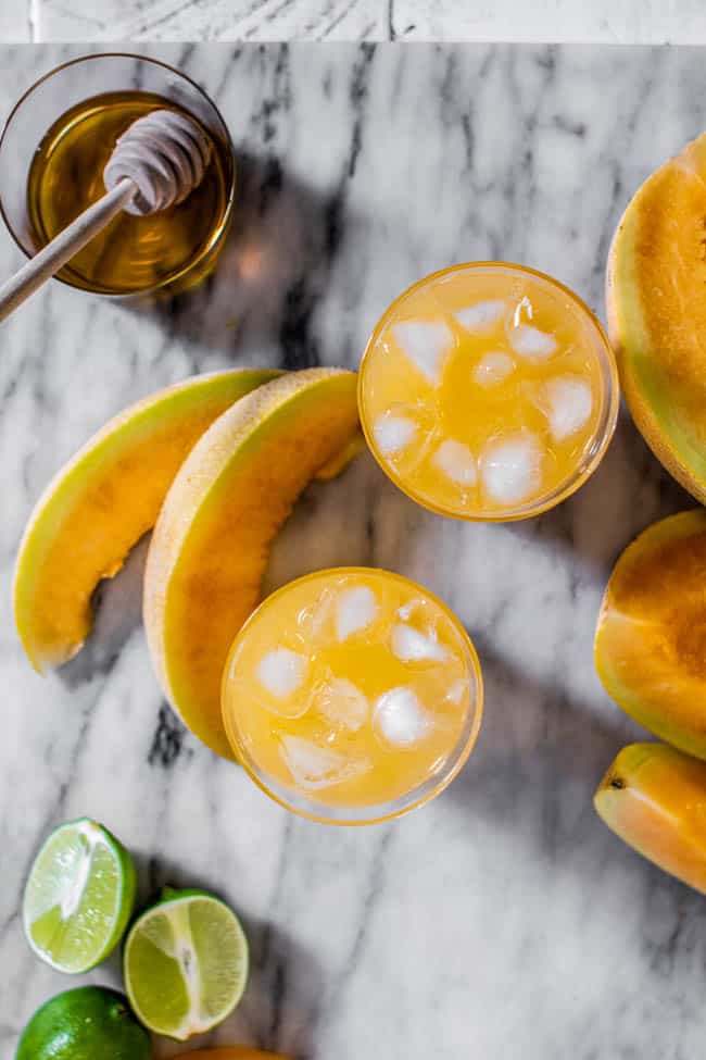 Two glasses of Cantaloupe Agua Fresca with ice on a marble board with lime halves and cantaloupe wedges.