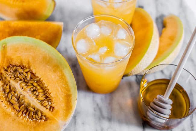 A glass of our Cantaloupe Agua Fresca recipe next to half a cantaloupe and a jar of honey.