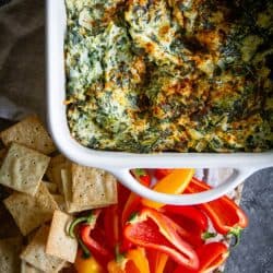 A baking pan fresh out of the oven filled with A Classic Artichoke and Spinach Dip Recipe. There are crackers and mini bell peppers for dipping.