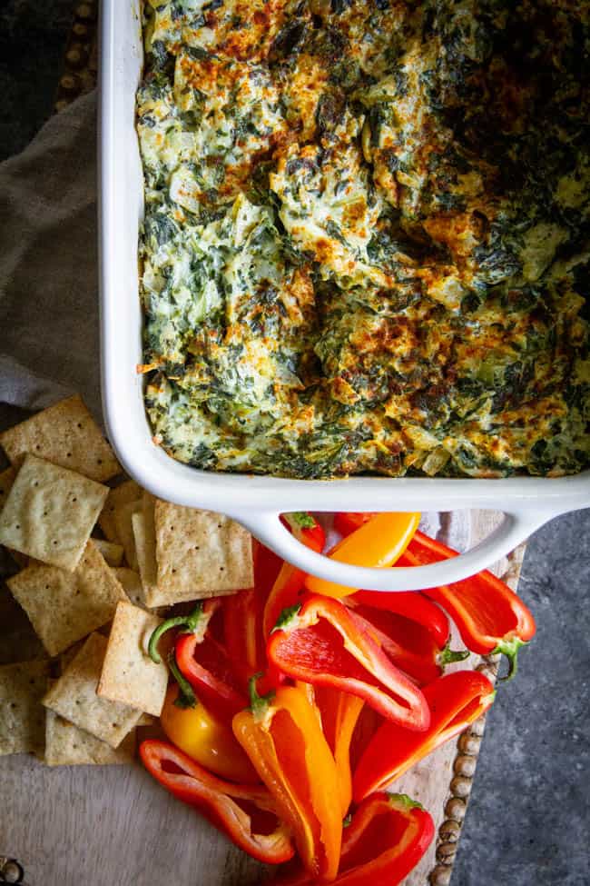 A baking pan fresh out of the oven filled with A Classic Artichoke and Spinach Dip Recipe. There are crackers and mini bell peppers for dipping.