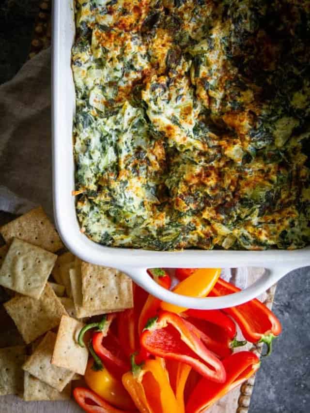 A baking pan fresh out of the oven filled with A Classic Artichoke and Spinach Dip Recipe. There are crackers and mini bell peppers for dipping.
