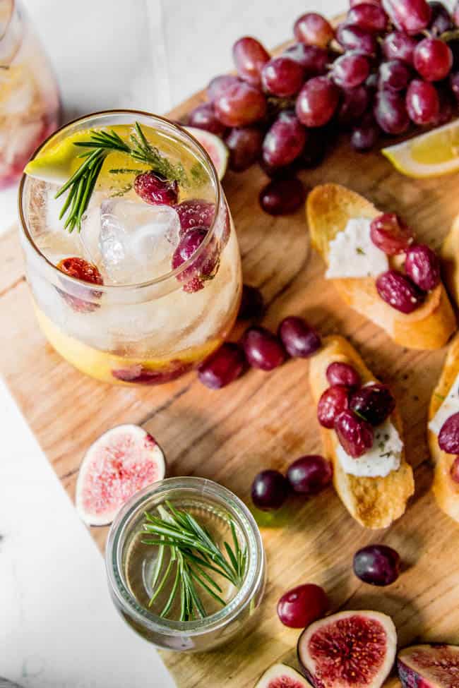  Rosemary and Roasted Grape Wine Cocktail on a serving board with crostini that has a slice of cheese and roasted grapes on top. 