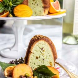A slice of Pistachio and Moscato Bundt Cake on a plate with basil leaves, stone fruit slices, and citrus wheels.