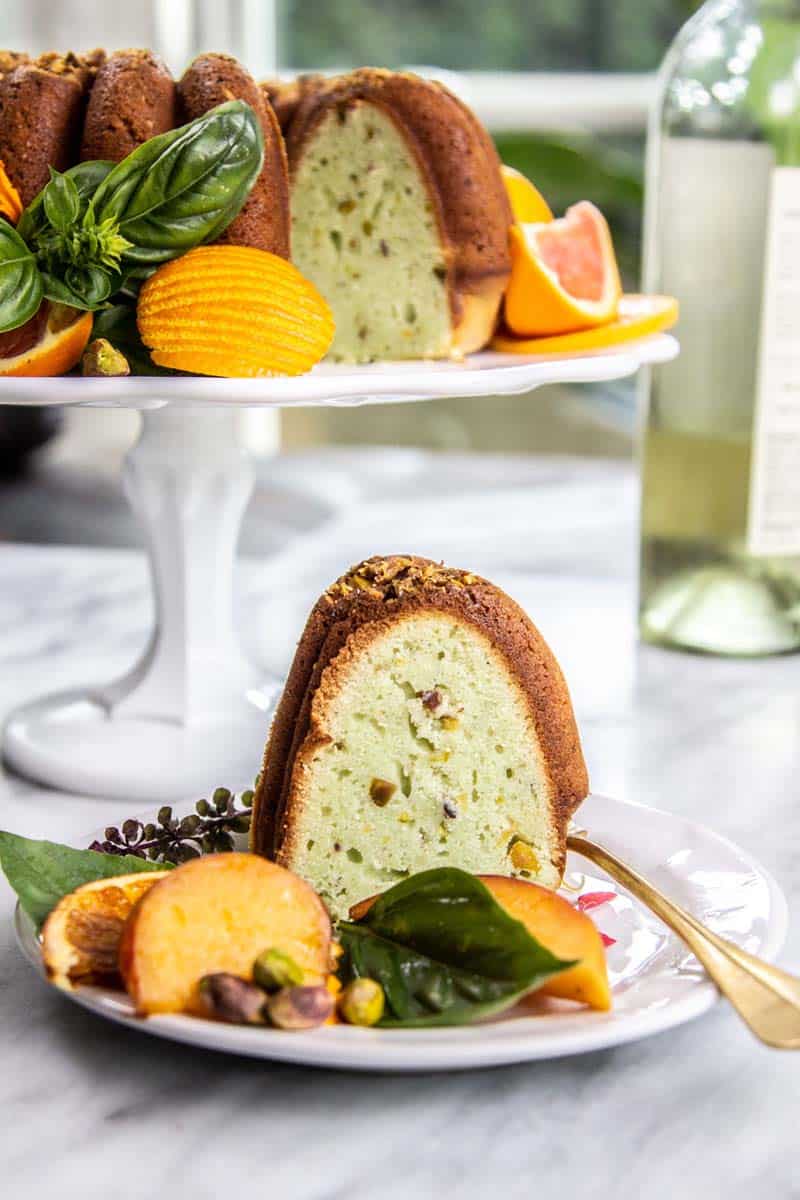 A slice of Pistachio and Moscato Bundt Cake on a plate with basil leaves, stone fruit slices, and citrus wheels.