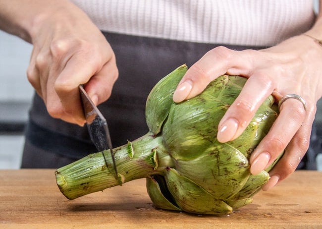 Woman holding an artichoke on a cutting board and using a knife to cut off the stem