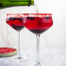 Coupe glasses with a red cockytail and a red rim. Saprkling wine is being poured into the glass.
