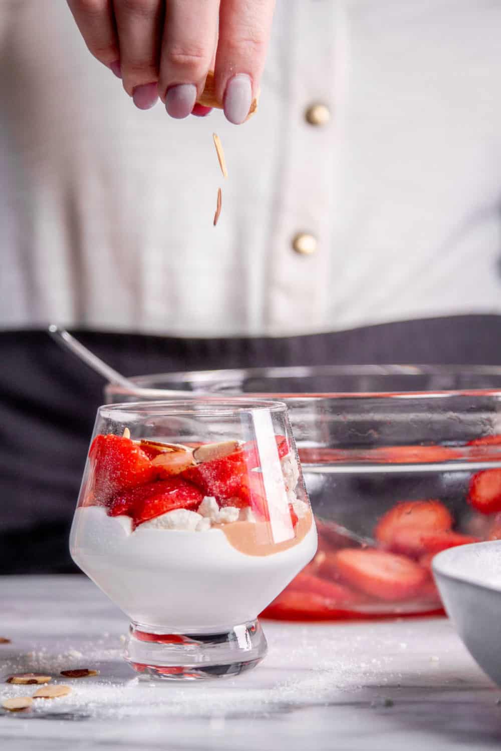 Adding toasted almonds into a glass with whipped cream, strawberry curd, meringues, and fresh strawberries.