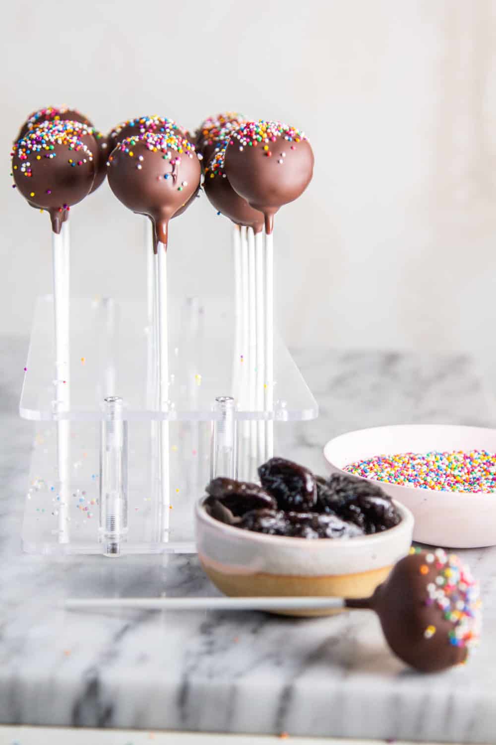 A bowl of prunes and a bowl of sprinkles on a marble slab next to a cake pop stand filled with sprinkle covered chocolate cake pops.