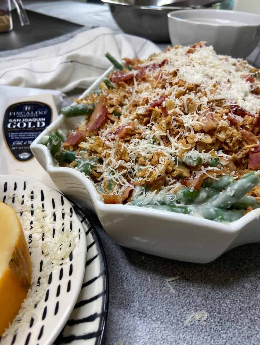Green bean casserole in a white bowl topped with crunchy onions, bacon and cheese