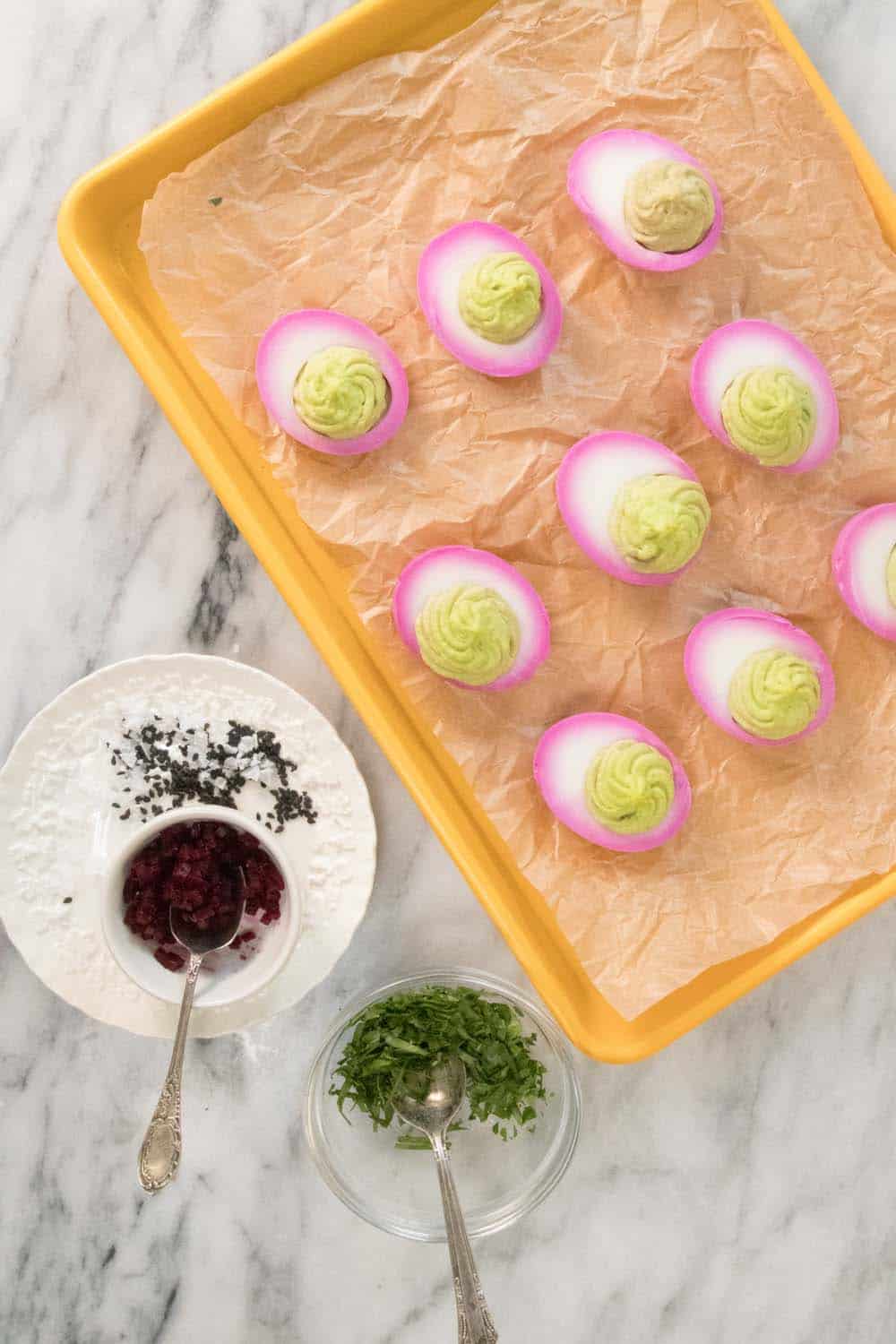 A sheet pan with deviled eggs ready to be garnished. The deviled eggs have pink rings around the outside of the egg white from picked beet juice and a light green filling.