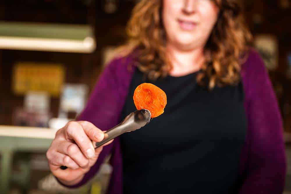 A woman holding a dried apricot with serving tongs for us to try.