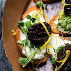 A crostini topped with red wine spiced prunes, salted ricotta, sprouts, lemon zest.