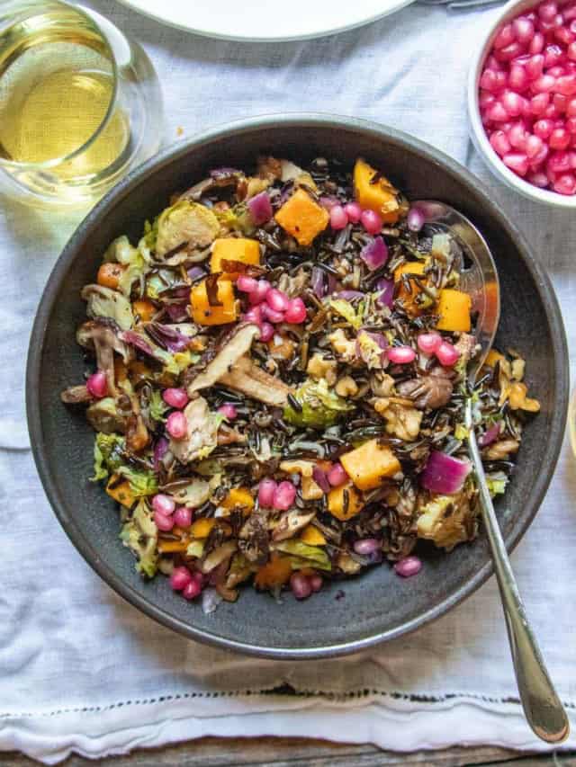 A serving bowl of Veggie Loaded Wild Rice Stuffing next to a glass of wine and a small bowl of pomegranate arils.